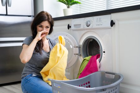Pictured is a women holding her nose while pulling out a yellow towel from a front-loading washing machine. There is a green, dark pink, and light pink towel hanging out of the front of the washer. The woman is crouching behind a grey laundry basket. The scene appears to be in a kitchen with a refrigerator in the background.