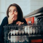 Woman leaning down next to her open dishwasher. She is closing her nose and looking away seemingly because of the dishwasher odor
