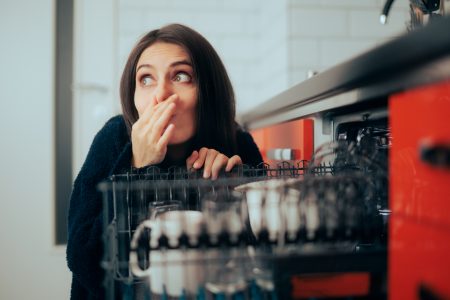 Woman leaning down next to her open dishwasher. She is closing her nose and looking away seemingly because of the dishwasher odor