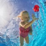 A young girl is swimming under the water in a swimming pool. She is reaching for a red flower.