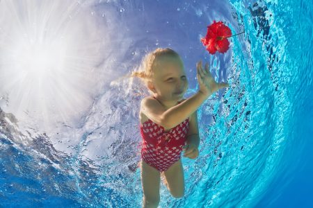 A young girl is swimming under the water in a swimming pool. She is reaching for a red flower.